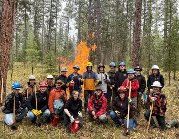 Group of students stand in front of burning pile in a forest